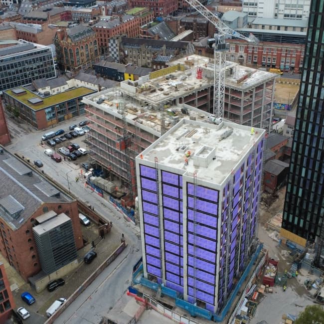 1NQ, Tariff Street, Manchester: aerial drone shot of the scheme mid-construction, completed core wrapped in violet Tyvek breather membrane, tower crane on top, surrounded by the red-brick mills and modern apartment blocks of the Northern Quarter rooftops.