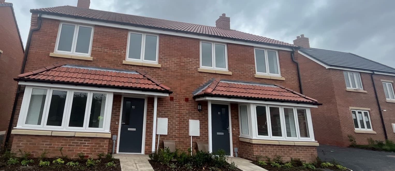 Aviation Lane, Burton-upon-Trent: a pair of completed red-brick three-bedroom semi-detached homes at numbers 20 and 21, terracotta-tiled pitched roofs and matching tile-roofed porches above charcoal front doors, white casement windows with stone lintels, shared paved driveway. The wider Midland Heart development of identical brick-and-tile pairs is visible on the right.