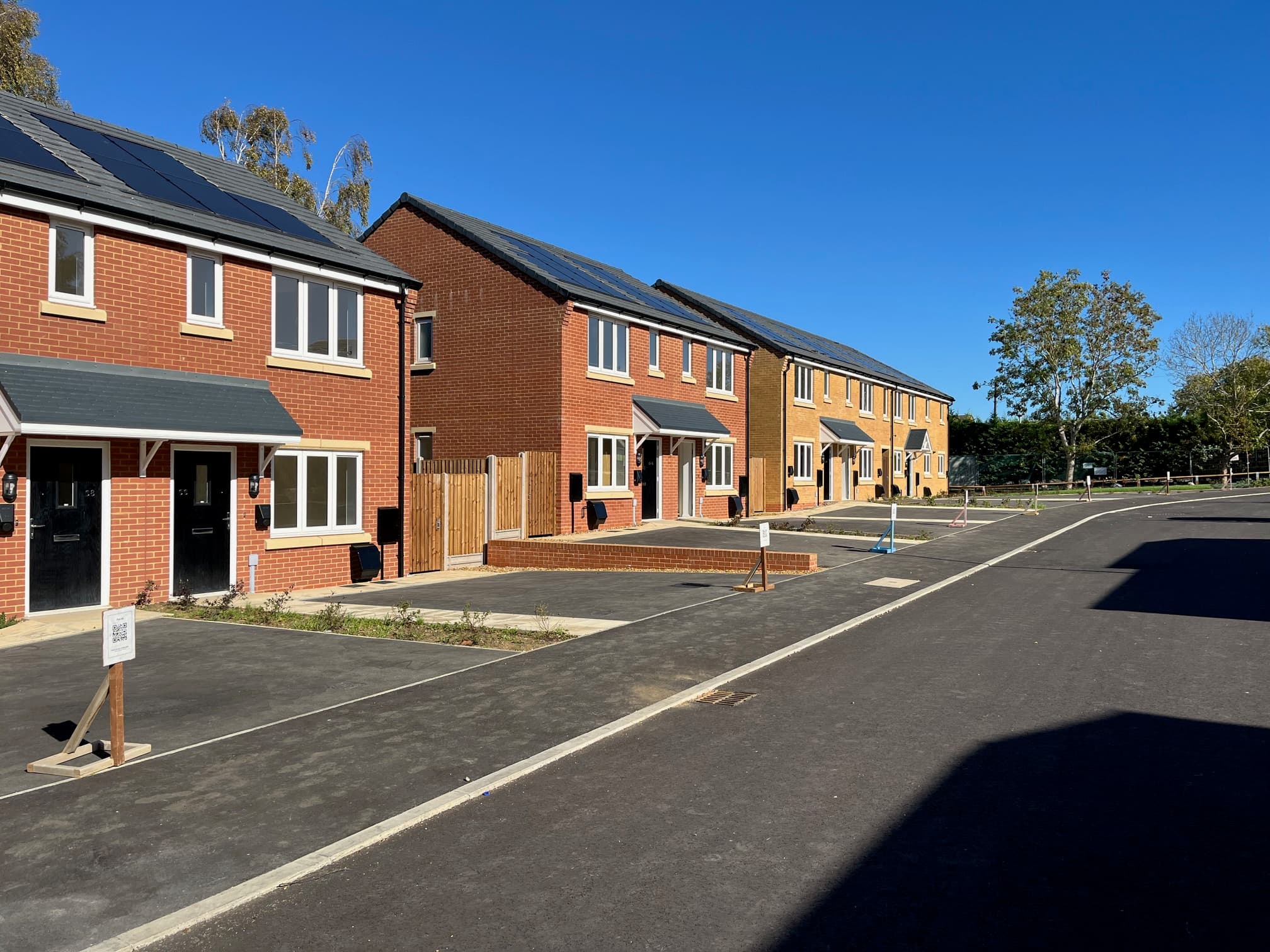Central Avenue, Northampton: streetscape of red- and yellow-brick semi-detached and detached homes with PV-clad pitched roofs running along a freshly tarmacked road, mature woodland visible in the background