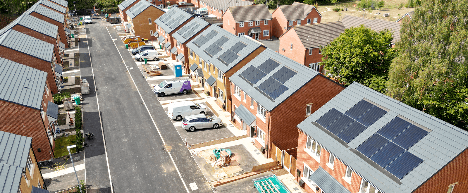 Central Avenue, Northampton: aerial drone view down the new residential street, terraced rows of yellow- and red-brick homes with PV-clad slate-grey pitched roofs, residents' cars and a contractor van parked along the kerb, mature trees ringing the site