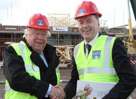 Chestnut Grange handover moment: two men in red Trent & Dove hard hats and hi-vis vests shaking hands at the construction site, scaffolding and Bullock Construction site hoarding visible behind, holding a 'Retirement living' brochure.