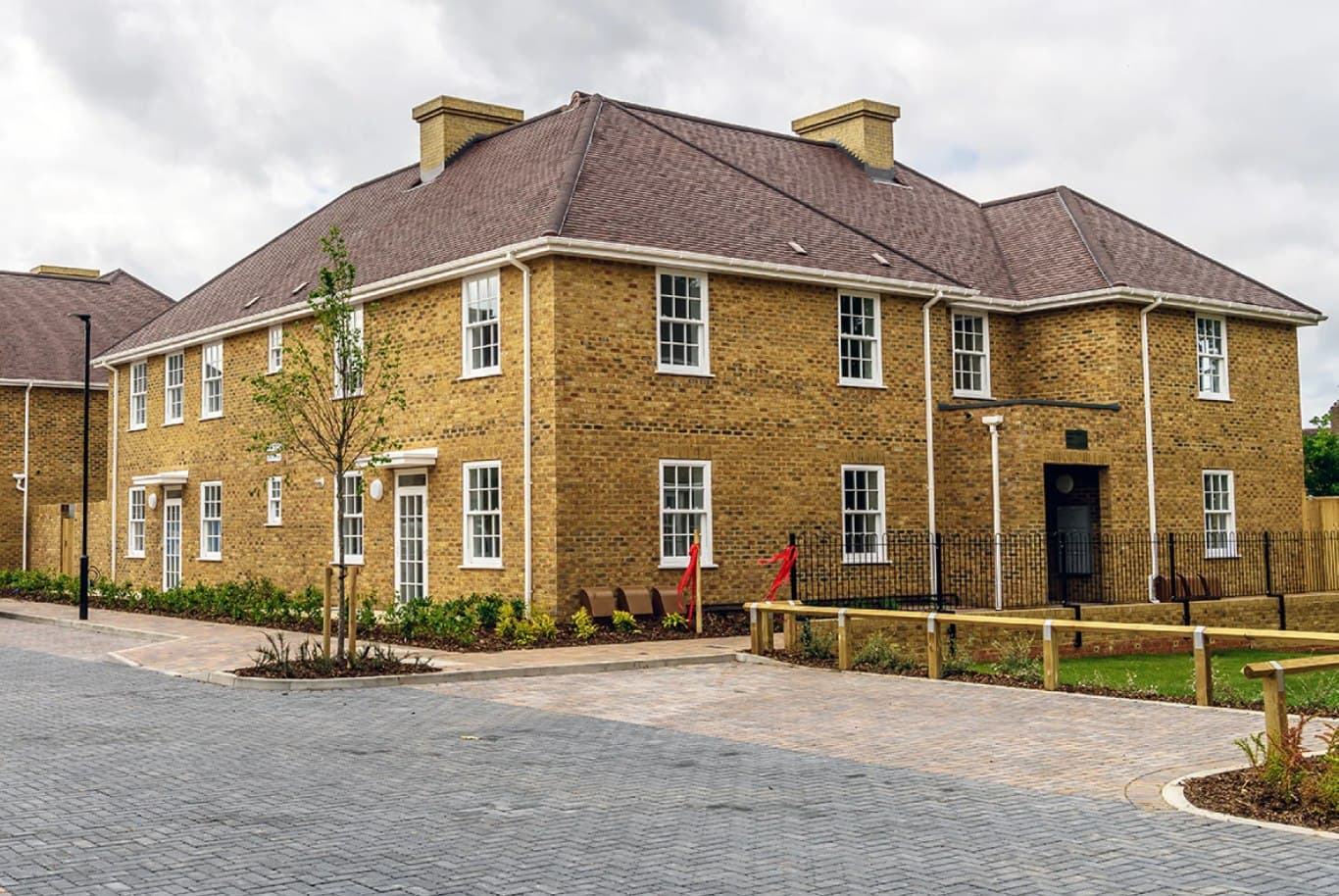 Haig Homes, Morden: traditional yellow-stock-brick apartment block with a hipped tiled roof and chimneys around a newly block-paved courtyard with timber benches and planting, Haig Housing scheme for the armed-forces community