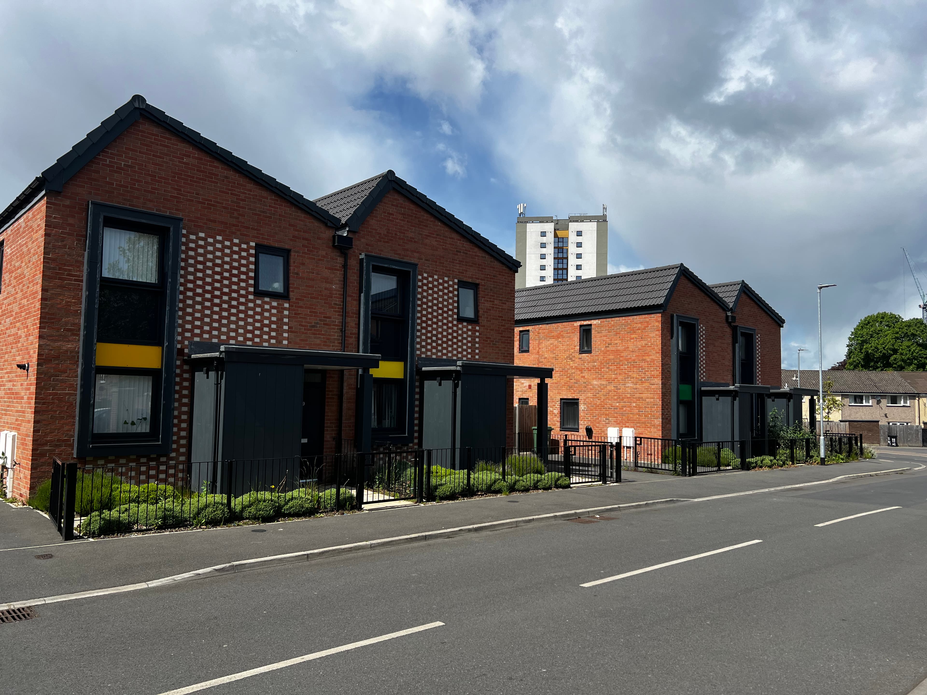 Leeds Meynell, Holbeck: two completed timber-frame two-storey houses with red and dark-brown brick facades, decorative perforated brickwork at the gable apex, dark-framed windows, low railings and planted hedging at the kerb. A residential tower block from the wider Holbeck regeneration zone is visible in the background.