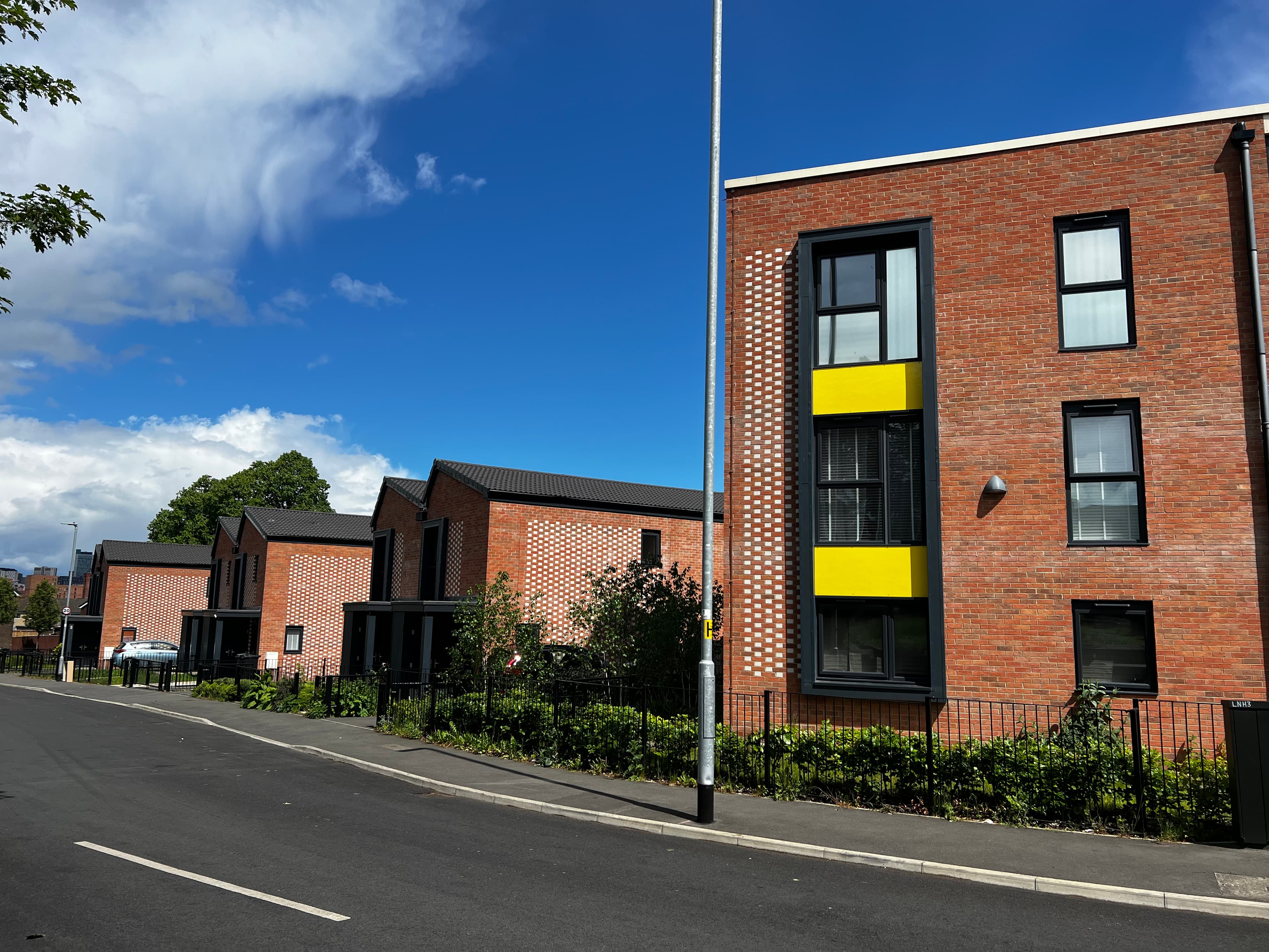 Leeds Meynell, Holbeck: three-storey one-bedroom apartment block with red brick walls, recessed yellow accent panels framing the upper windows, decorative perforated brickwork running up the lift core, framed by clear blue sky.