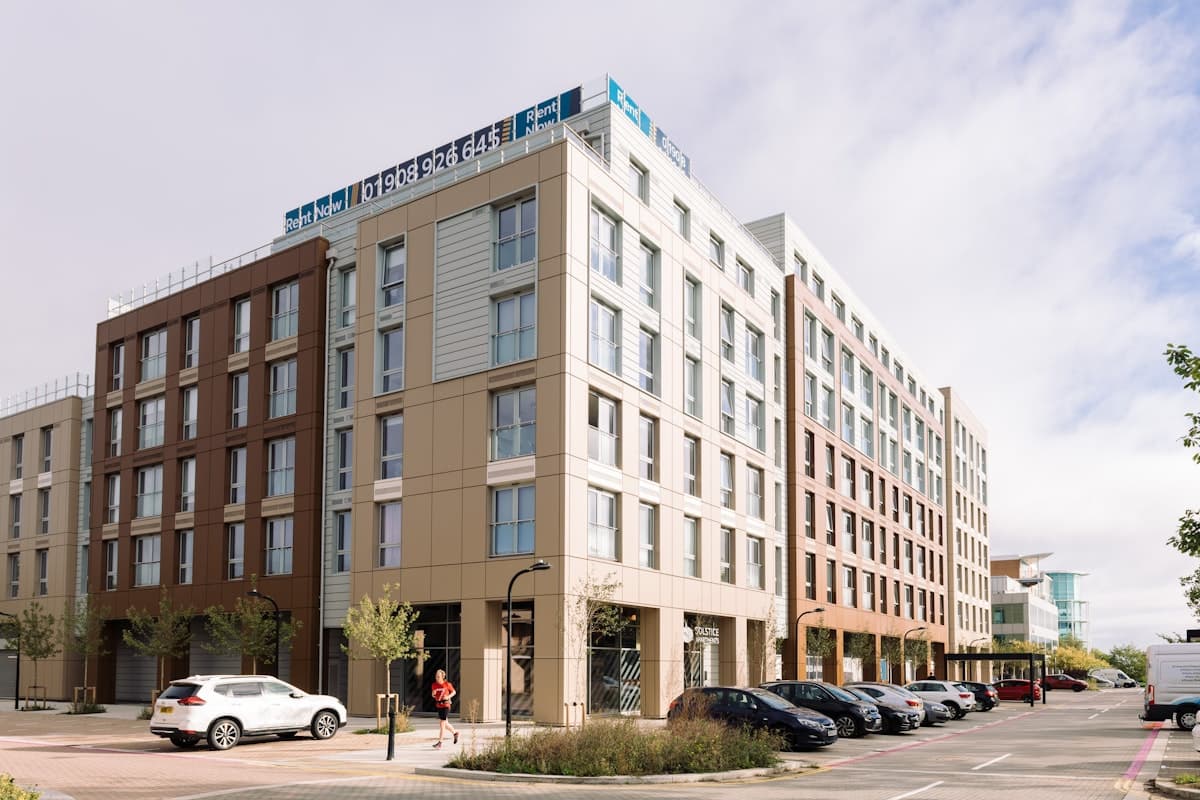 Solstice Apartments, Milton Keynes: completed eight-storey BTR block on Silbury Boulevard in pale and warm-tan cladding with grid windows, surface car park in the foreground and rooftop "Rent Now" signage, Grainger plc scheme