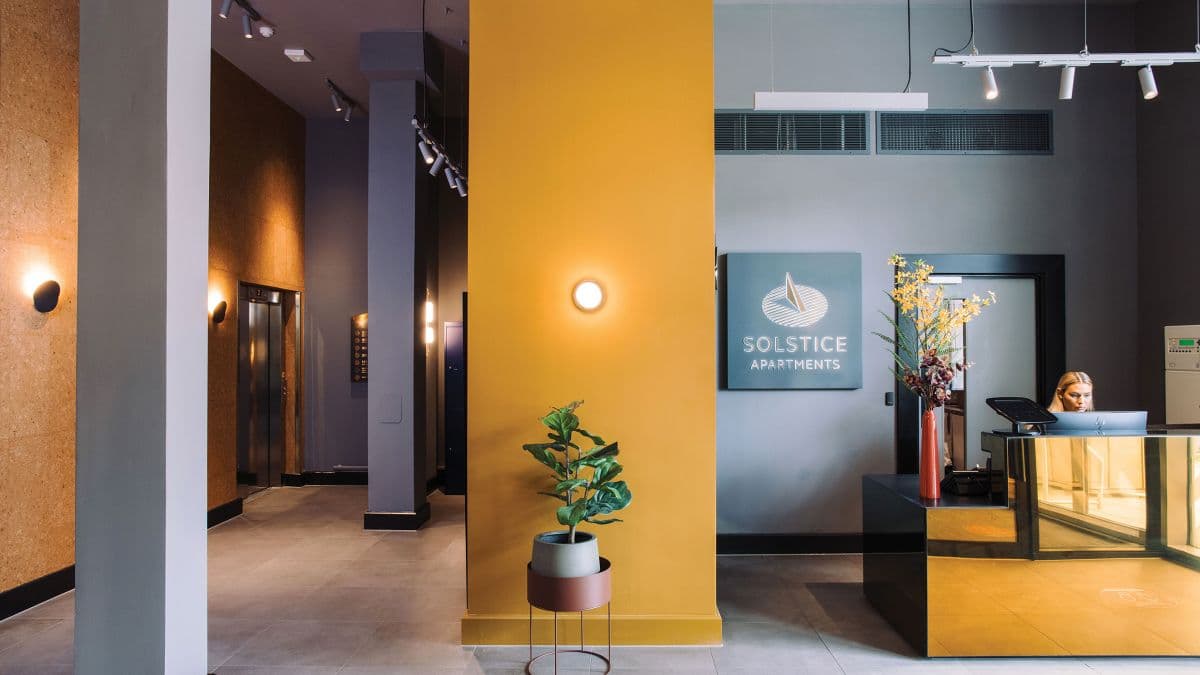 Solstice Apartments, Milton Keynes: residents' lobby with mustard accent wall, branded "Solstice Apartments" sign behind a brass-clad concierge desk, lift bay and oak-cork wall to the side