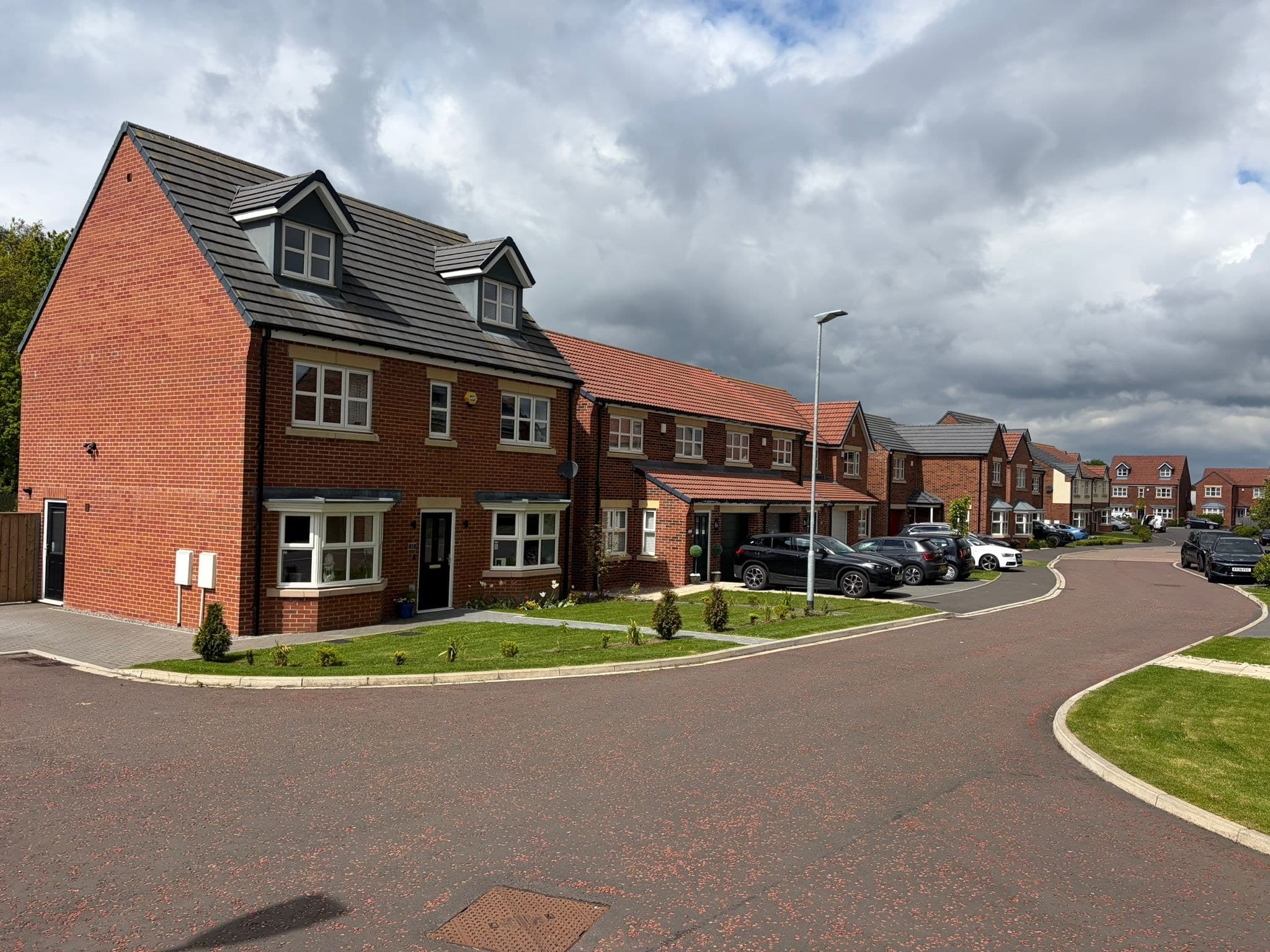 Spennymoor (Hartwall) Phase 2: completed two- and three-storey red-brick homes with grey pitched roofs and dormer windows on a newly-laid residential close, parked cars and landscaped front gardens visible, Tarras Park Properties affordable scheme.