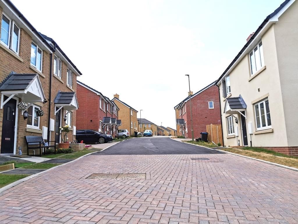 Springfield Oval, Witney: residential street view at handover looking down a brick-paved road bordered by mixed cream-render and red-brick two-storey houses with slate pitched roofs and gabled porch canopies, parked cars and bench seating along the kerb