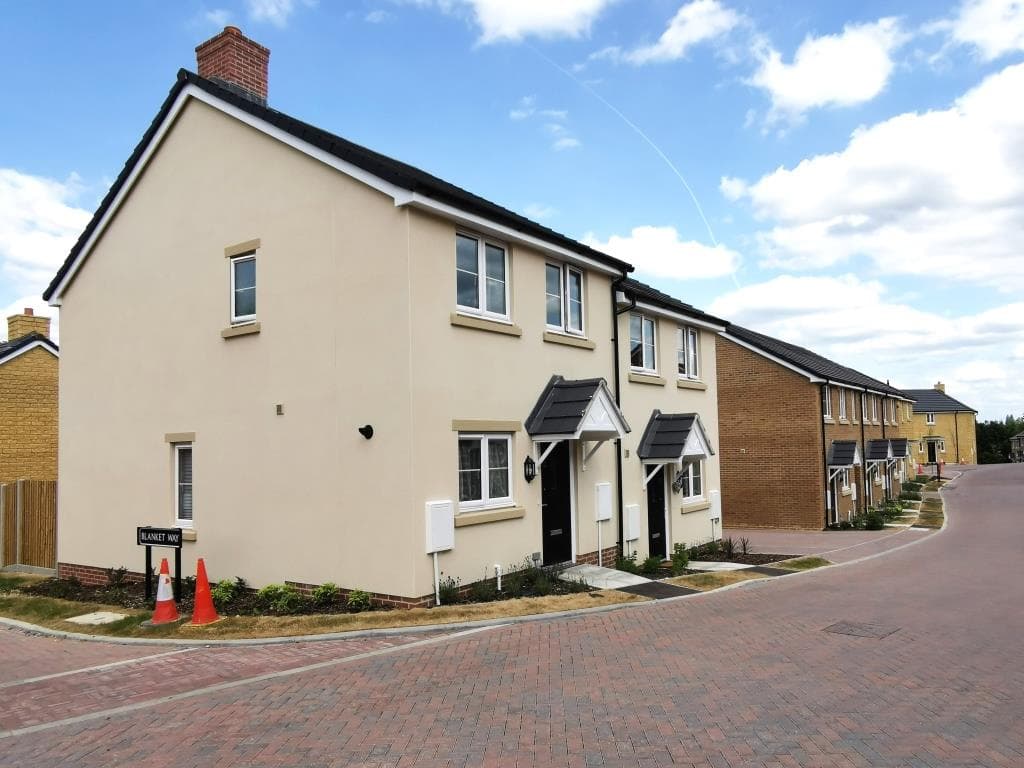 Springfield Oval, Witney: gable-end view of a cream-rendered two-storey semi-detached house with red-brick chimney, slate pitched roof, traditional canopy porches and stone-effect window surrounds, set on the corner of Blanket Way at handover, with the brick-paved estate road and the rest of the development running off to the right