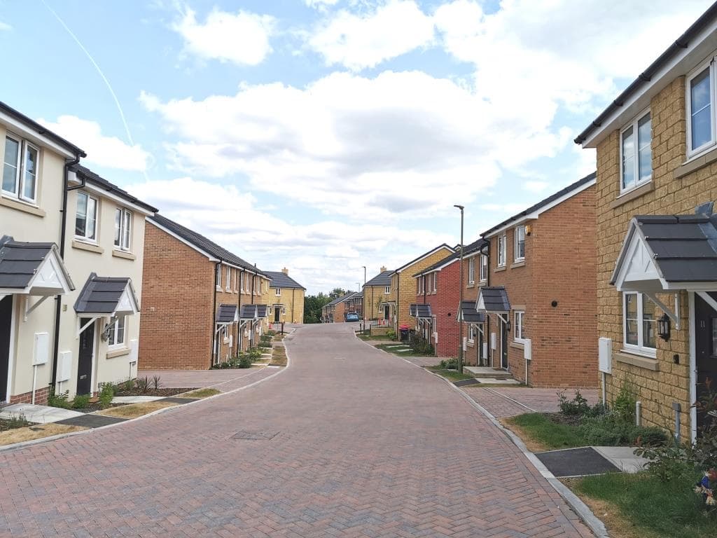 Springfield Oval, Witney: view down a new block-paved estate street at handover, terraces of two-storey cream-render and red-brick houses with slate pitched roofs and traditional gabled porch canopies on both sides, young landscaping just bedded in