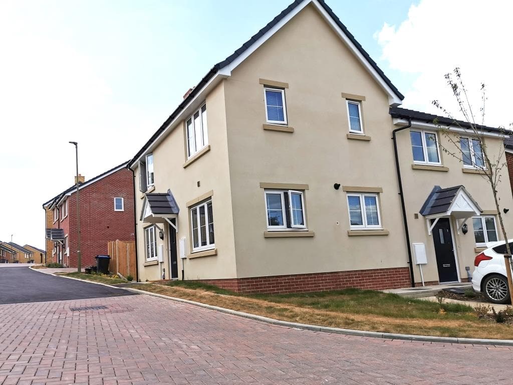 Springfield Oval, Witney: corner three-bay cream-rendered detached house with stone-look quoins, traditional window surrounds and a steep slate pitched roof, with adjacent dwellings in mixed render and red-brick palettes and a young street tree on the freshly-laid grass strip