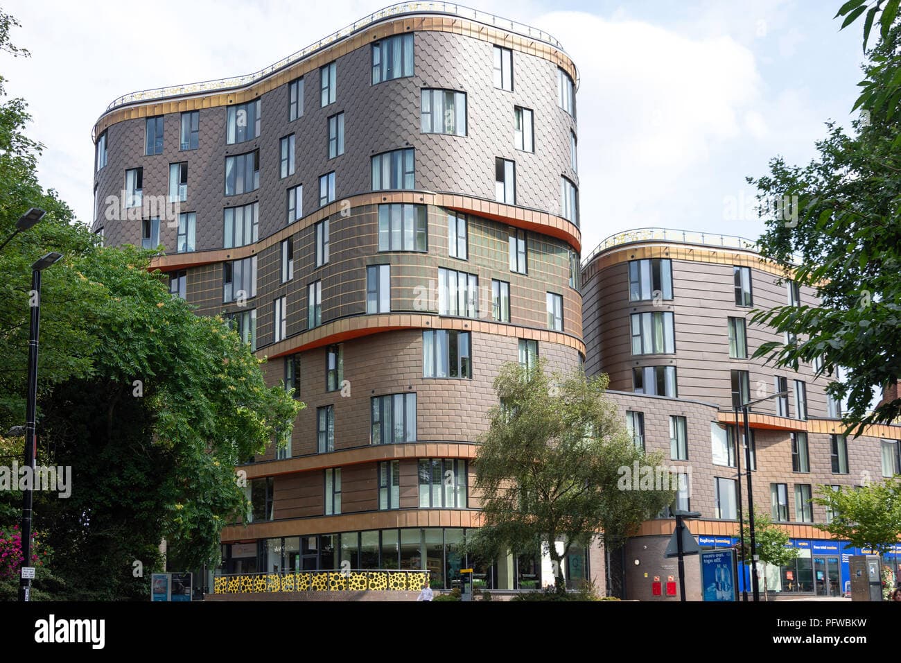 The Fold, Sidcup: a sculpted residential block in copper-brown tile cladding, multiple curved volumes stacking around a central core with white-framed windows, trees and street furniture in the foreground, completed scheme on Sidcup High Street, London Borough of Bexley.