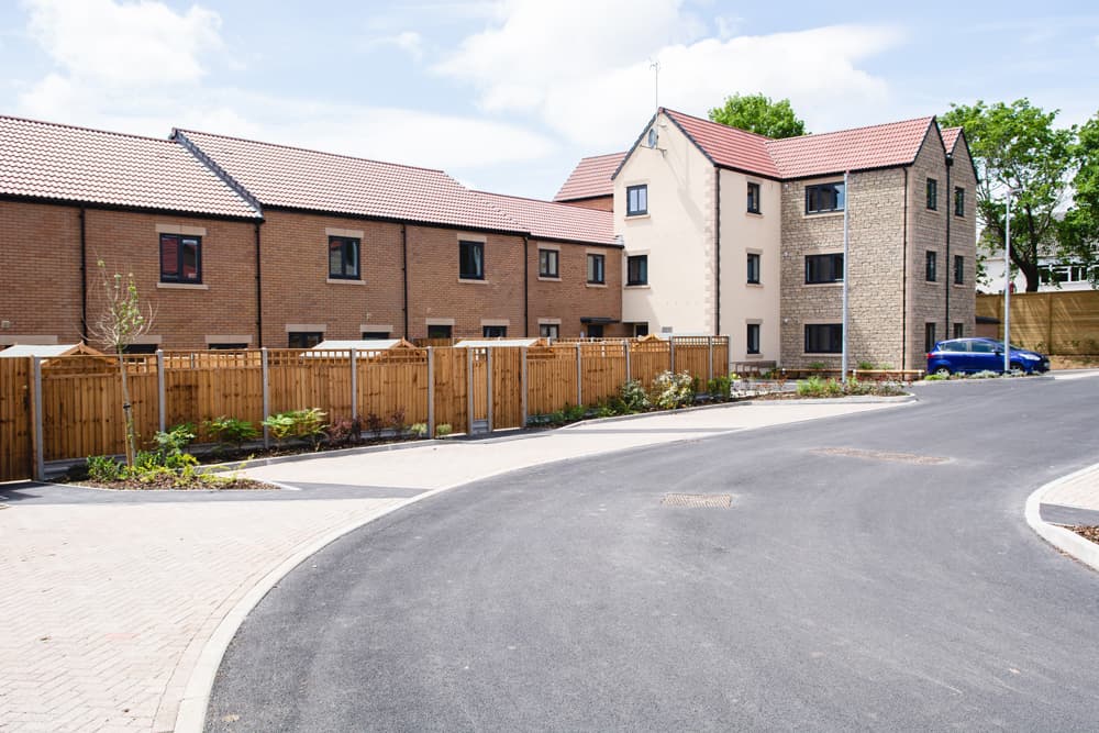 The Moorings, Pucklechurch: site view along a new estate road past timber-fenced rear gardens, terraces of brick and rendered homes beyond