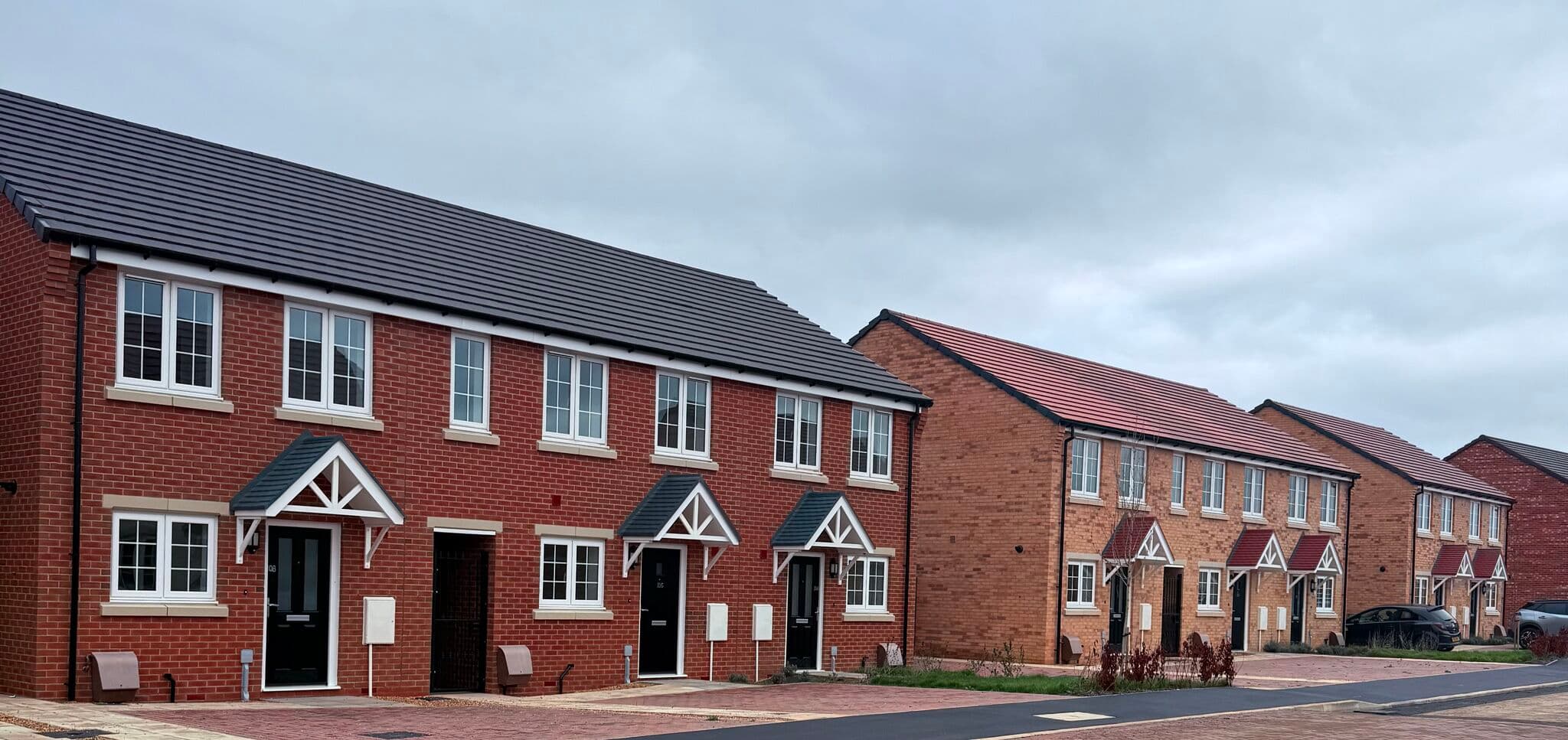 Wisbech Road, March: completed terrace of red-brick two-storey homes with grey pitched roofs and white-trimmed pitched porch hoods, paving-block driveway running across the foreground, Sage Homes affordable scheme